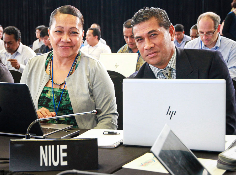 Niue's Associate Minister for Natural Resources, Hon. Esa Sharon-Mona Ainuu, left, at WCPFC16, sitting at a table in a meeting room, next to an unnamed man. Country label Niue in front of them, and they have laptop computers open in front of them, and a microphone.