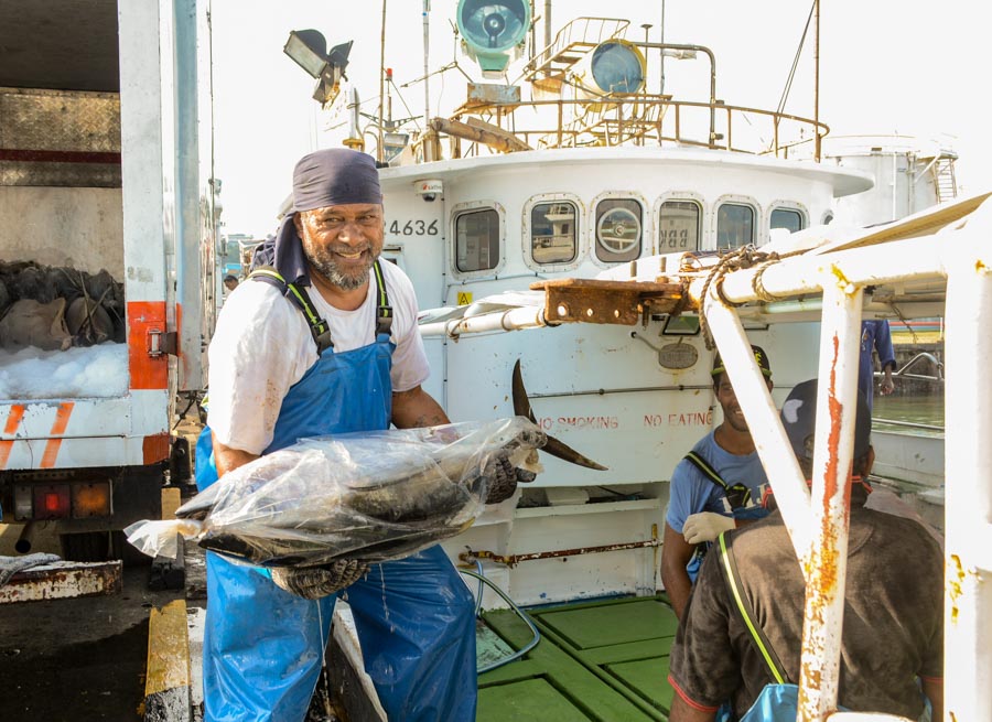 Smiling man wearing bandana on head and large plastic apron holds whole bagged tuna on wharf. Fishing boat in background.