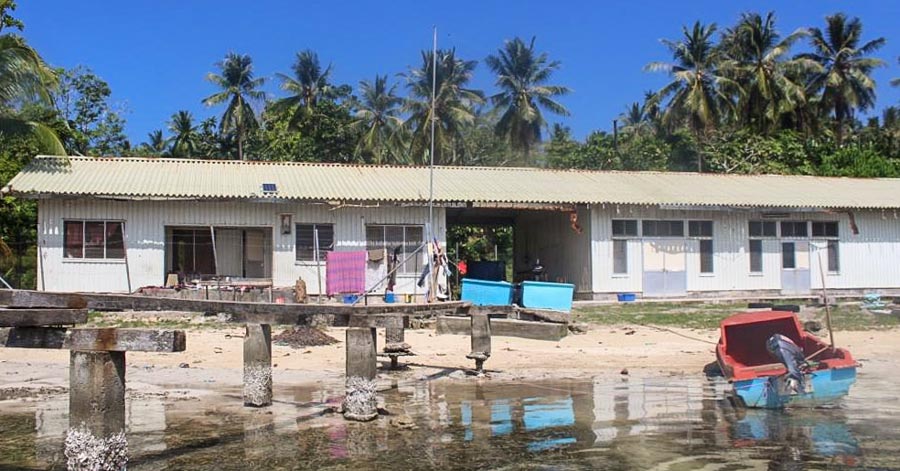 A long, low building with a corrugated metal roof stands near the water. Two large plastic boxes stand in front. In the shallow water, a dingy with an outboard motor is at anchor.