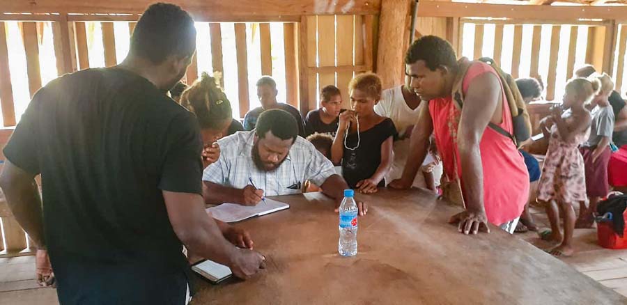 Man sits at table writing in book while other tribal landowner applicants look on, Bina Harbour beneficiary mapping, Solomon Islands