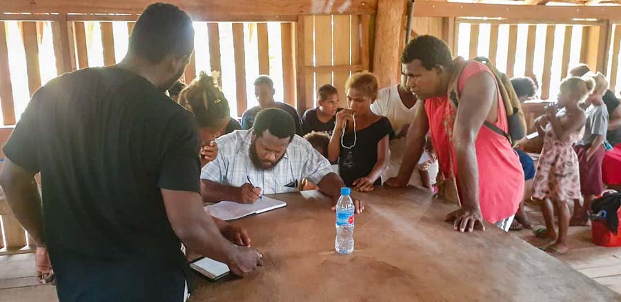 Man sits at table writing in book while other tribal landowner applicants look on, Bina Harbour beneficiary mapping, Solomon Islands