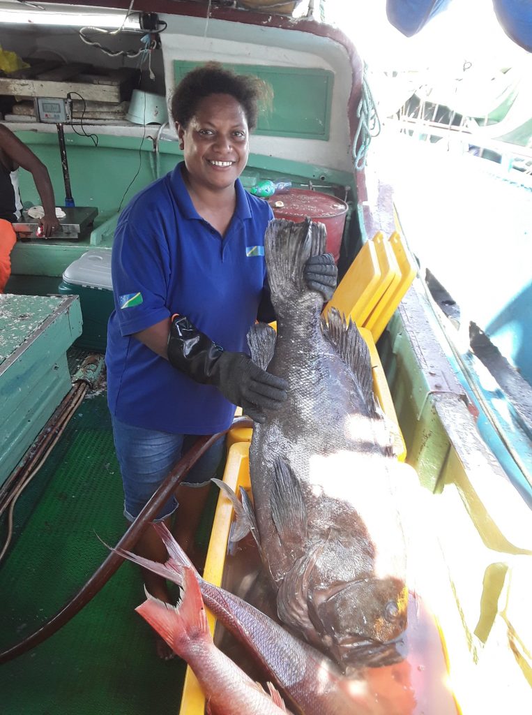 A worker with gloves holds a large fish in a bucket on the deck of a ship