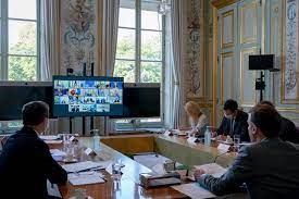 Officials seated around a desk looking at a Zoom meeting grid of faces on a monitor