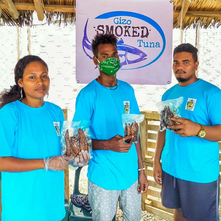 Young woman and 2 young men in turquoise t-shirts each hold a vacuum-packed packet of smoked fish in front of sign that read Gizo Smoked Tuna.