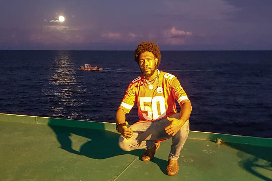 Jeromy Wateoli, young man of Solomon Islands, crouches on a flat metal surface, giving thumbs up. Behind him is the ocean and the moon.