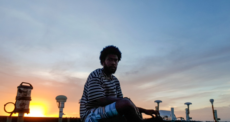 Man sits on boat structure, silhouetted against setting sun