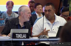Two men seriously talk at a desk labeled Tokelau with a microphone in front of them